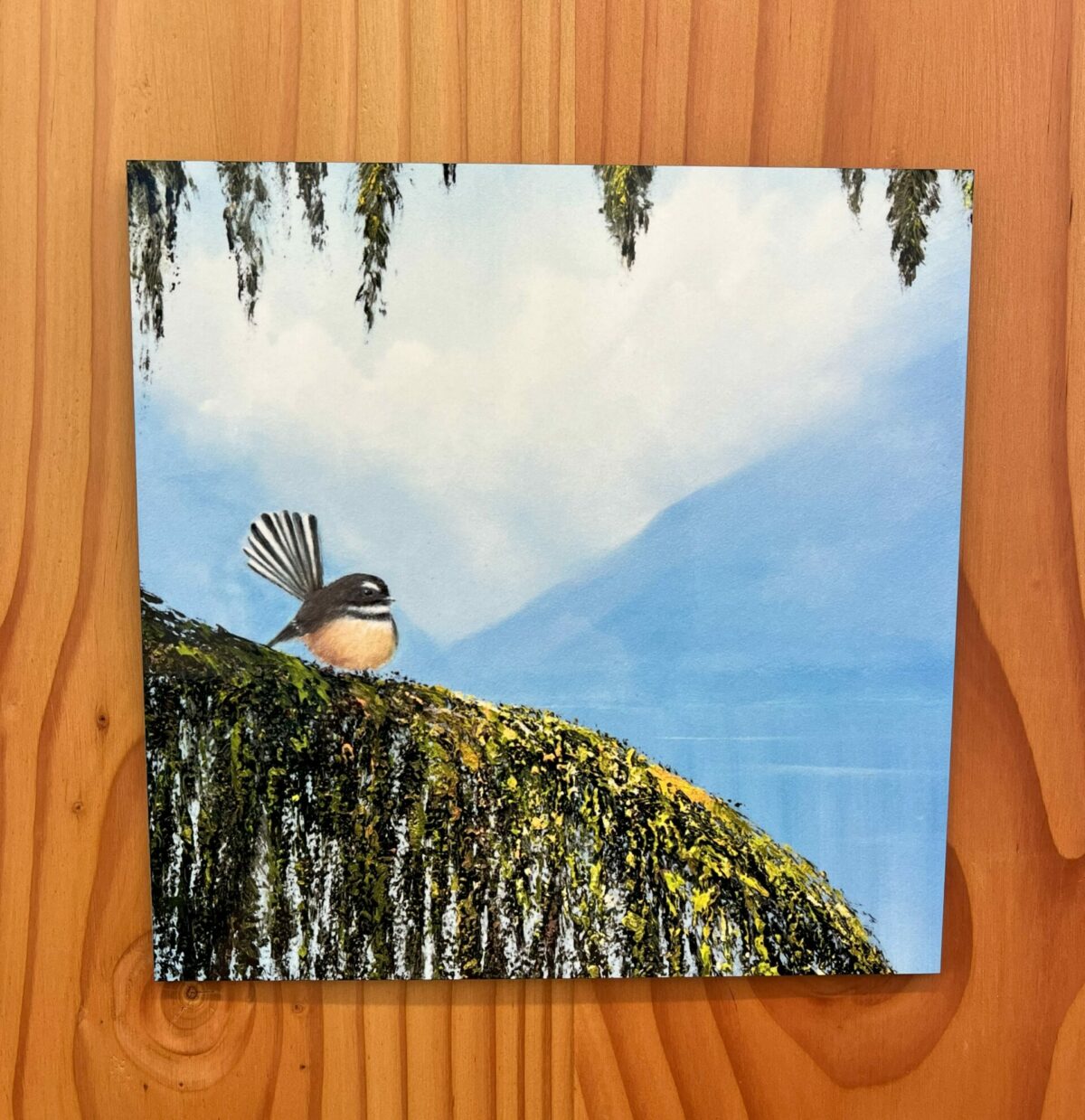 Fantail Wall Tile features a depiction of a small bird with a fanned tail perched on a mossy branch, set against a backdrop of blue sky and distant mountains.