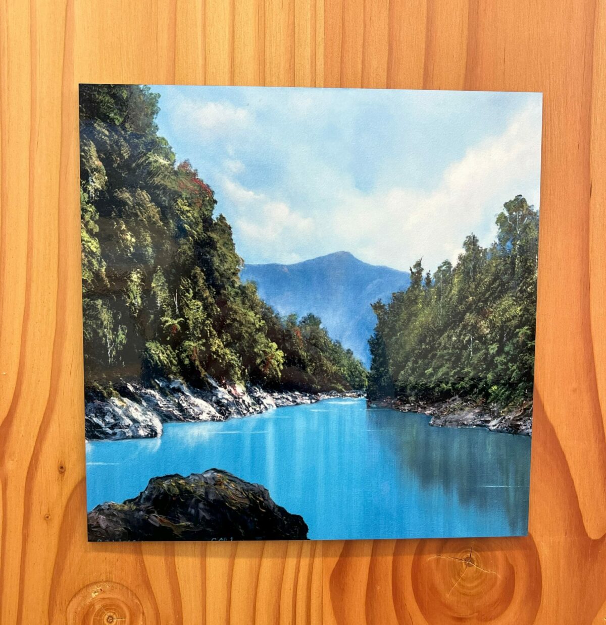 A painting of Hokitika Gorge, depicting a serene river flowing between tree-covered banks under a blue sky with distant mountains.