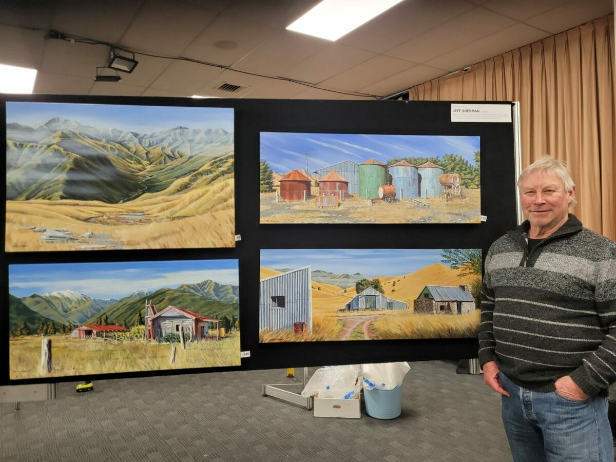 A man stands beside a display of four "Into the Tuke" landscape paintings, showcasing rural scenes with mountains and fields.