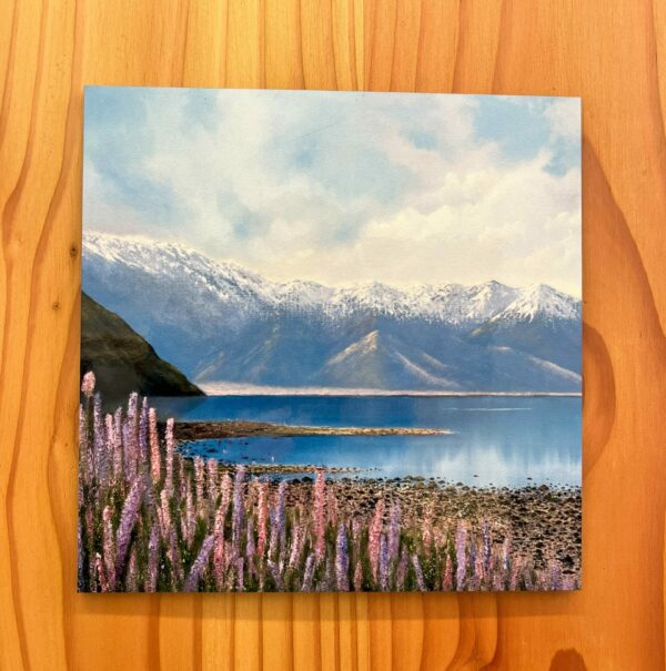 Printed Wall Tile of Lake Tekapo: Features a peaceful lake with remote snow-capped mountains, a partly cloudy sky overhead, and purple flowers in the foreground, all set against a wooden surface.