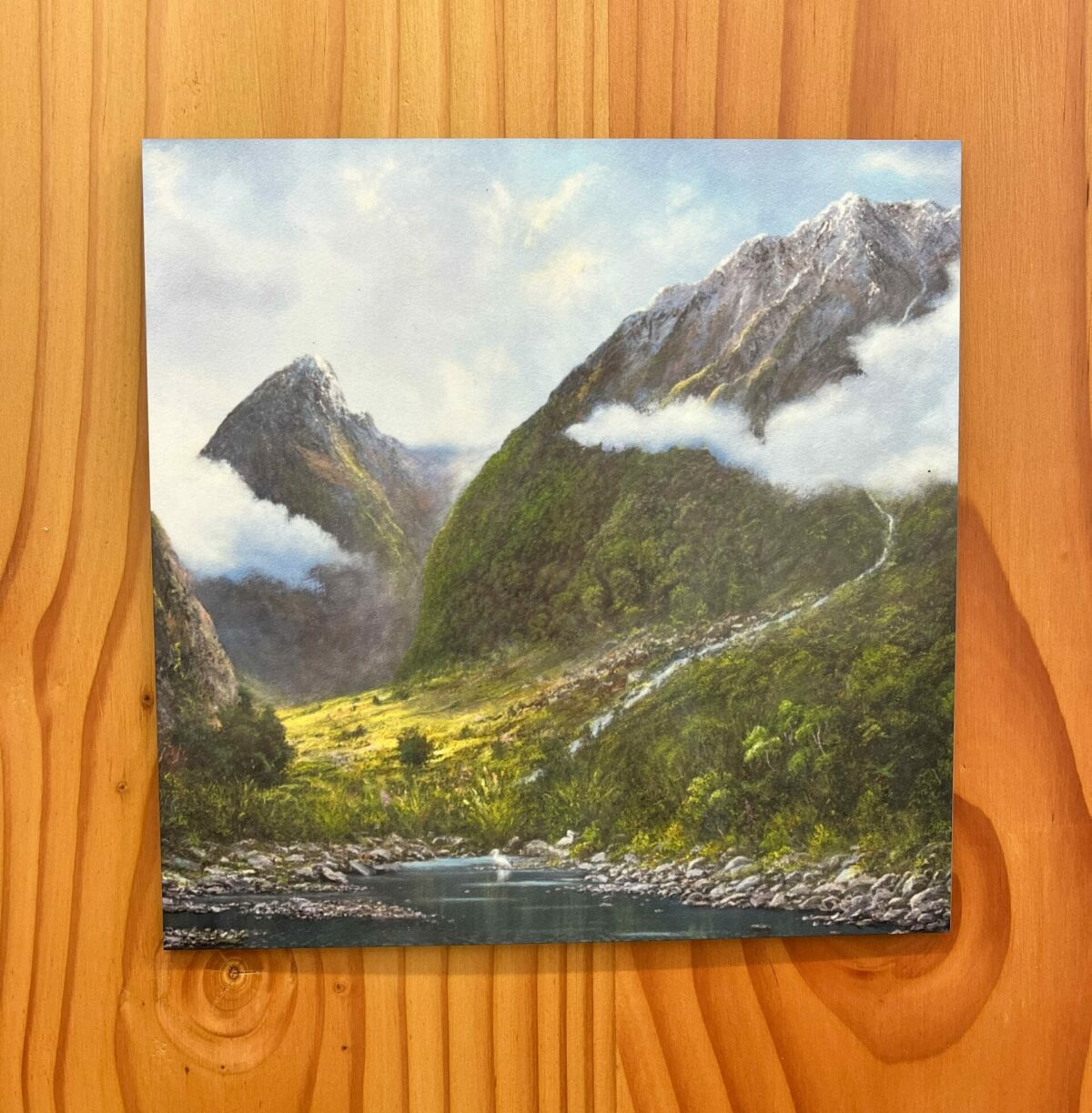 A Printed Wall Tile of Mount Talbot featuring a mountain landscape with two peaks, a flowing river, forested slopes, and some low clouds set against a clear sky, beautifully displayed on a wooden wall.