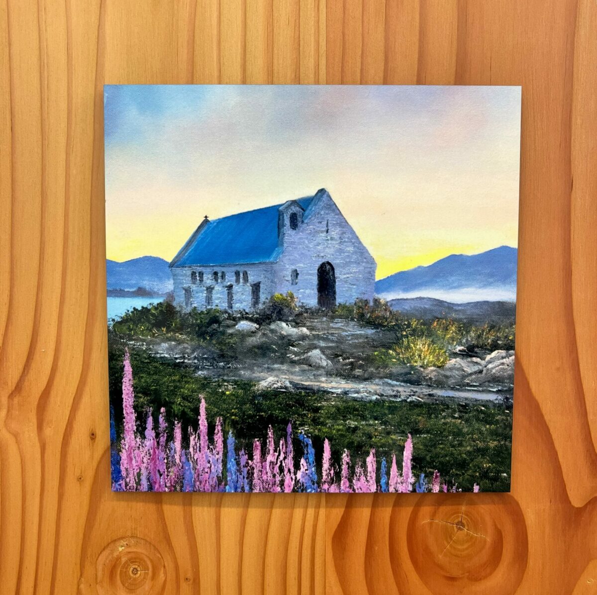 The Church of the Good Shepherd Wall Tile features an image of a small stone building with a blue roof, surrounded by hilly landscapes and pink flowers in the foreground.