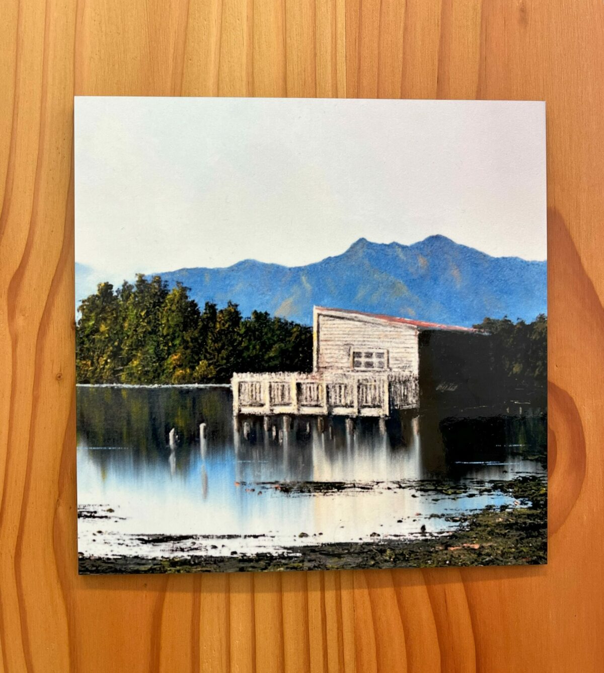 The Okarito Lagoon Boatshed wall tile features a wooden structure on stilts overlooking a tranquil body of water, with a backdrop of tree-lined mountains, all set against a wooden surface.