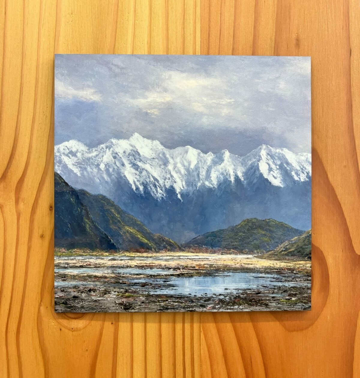 The Southern Alps Wall Tile features a painting that portrays snow-capped mountains, green hills, and a river beneath a cloudy sky, all set against a wooden backdrop.